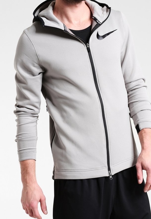 Homme portant un sweat à capuche zippé gris clair de Nike avec logo noir et pantalon noir, cadré au niveau du cou et du milieu des cuisses.