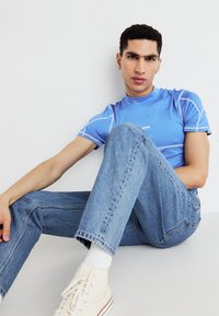 Blue short-sleeve athletic shirt with white seams, paired with light blue jeans and cream-colored canvas shoes, seated with legs crossed.