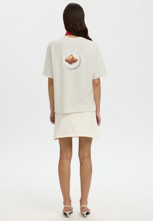 Kvinne iført en hvit kortermet topp med et croissantbilde på baksiden, kombinert med et matchende hvitt skjørt og beige hæler.