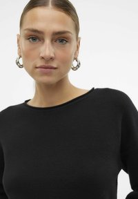 Pull noir texturé avec un col rond. Le modèle porte de grandes boucles d'oreilles en argent. L'arrière-plan est blanc uni.
