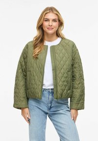 Jauna moteris vilki žalią siuvinėtą striukę, baltą marškinėlį ir šviesiai mėlynas džinsus, šypsosi ir stovi atsipalaidavusiomis rankomis šalia kūno.