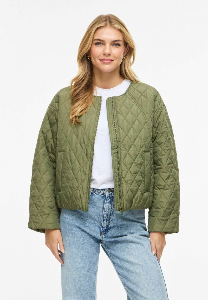 Jauna moteris vilki žalią siuvinėtą striukę, baltą marškinėlį ir šviesiai mėlynas džinsus, šypsosi ir stovi atsipalaidavusiomis rankomis šalia kūno.