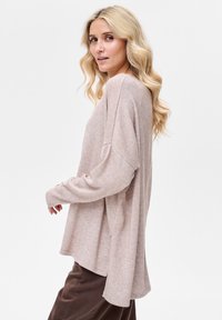 Jasnoróżowy, oversize'owy sweter z długimi rękawami, miękką fakturą i bocznymi rozcięciami; w połączeniu z brązowymi spodniami. Kobieta pozuje lekko odwrócona.