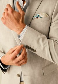 Veste de costume gris clair avec quatre boutons noirs sur le poignet, associée à une chemise blanche avec un col à motifs et un mouchoir décoratif.