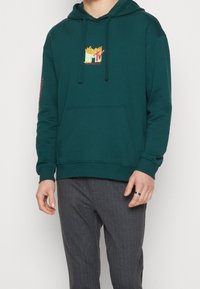 Sweat à capuche bleu sarcelle en coton, avec un grand logo MTV jaune et rouge sur la poitrine et du texte sur les manches. Modèle avec poche kangourou.