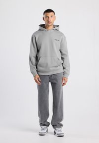 Uomo che indossa un hoodie grigio chiaro, jeans grigi larghi e sneakers bianche, in piedi di fronte a uno sfondo bianco semplice.