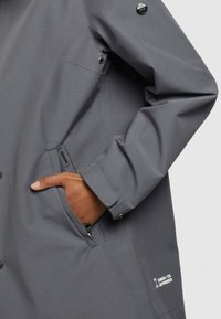 Ruka v kapse šedého saka se zipovou kapsou, knoflíkovou manžetou a malou značkovou nášivkou u dolního lemu.