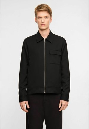 Ung mand iført en sort lynlåstrøje med en brystlomme og sorte bukser, stående foran en ensfarvet lys baggrund.