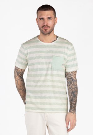 Uomo con capelli corti e tatuaggi su entrambe le braccia, indossa una maglietta a righe verdi chiare e bianche con taschino, in piedi davanti a uno sfondo bianco.