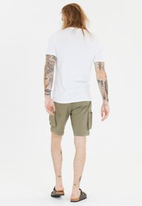 Wit katoenen t-shirt, olijfgroene cargoshorts met zijzakken en bruine instap schoenen. Tatoeages zichtbaar op armen en benen.