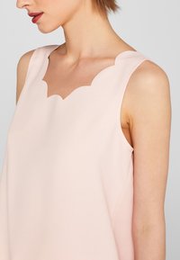 Kvinna som bär en ärmlös ljusrosa topp med en vågig halsringning, som visar hennes hals, axel och del av hennes ansikte i profil.