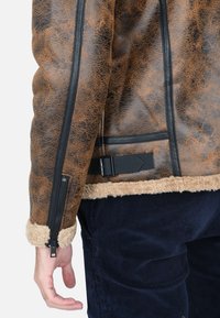 Giacca in eco-pelle marrone con foderatura in shearling, zip nera e dettagli con cinghie. Presenta una superficie testurizzata, tasca con zip e cuciture robuste.