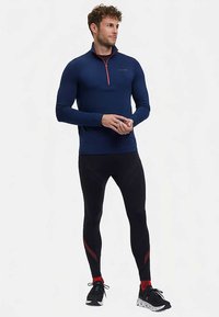Marineblauw atletisch shirt met lange mouwen en een rode rits, gecombineerd met zwarte strakke leggings met rode accenten. Zwarte sportschoenen maken de outfit compleet.