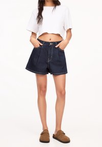 Shorts de mezclilla en azul oscuro con una cintura alta, con un cierre frontal de botones y dos bolsillos laterales. Camiseta blanca recortada y zapatos marrones tipo slip-on.