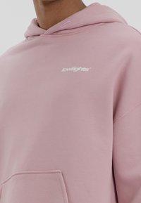 Gros plan sur une personne portant un sweat à capuche rose pastel avec le logo "lowlights" en blanc sur la poitrine gauche, une poche avant visible.