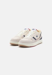 Beige suède en textiel sneakers met marineblauwe accenten, geperforeerde neus, en een witte rubberen zool. Kenmerkt een Lacoste-logo aan de zijkant.