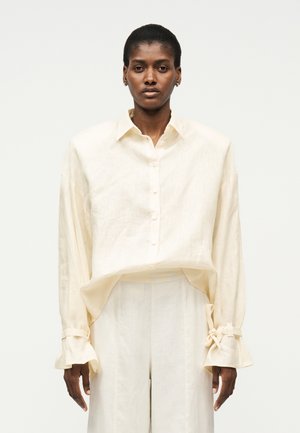 Personne noire portant un chemisier ample en lin de couleur crème avec devant boutonné et manches nouées, assorti à un pantalon large de même couleur, debout devant un fond blanc.