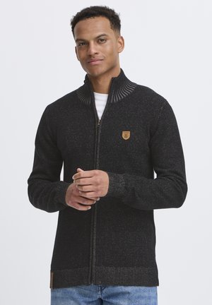 Sudadera negra con cremallera que presenta un cuello acanalado, una tela texturizada con detalles moteados y un pequeño parche con logo en el pecho.