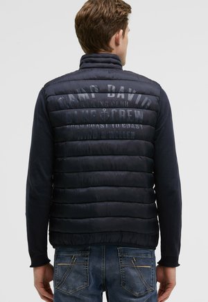 Man wearing navy blue quilted vest with "Camp David Blue Crew" text on back, dark long sleeves, and blue jeans, facing away.