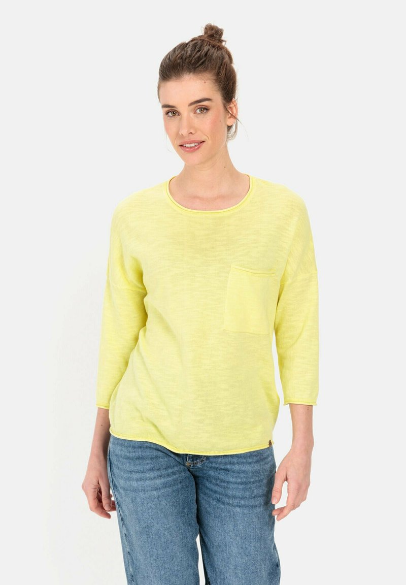 Gelbes leichtes Sweatshirt, Rundhals, drei Viertelärmel, lockere Passform, mit einer vorderen Tasche und subtiler Textur. Kombiniert mit blauen Jeans.
