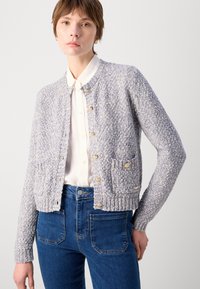 Textured light blue cardigan with gold buttons, featuring two front pockets and ribbed cuffs, worn over a white collared shirt and blue jeans.