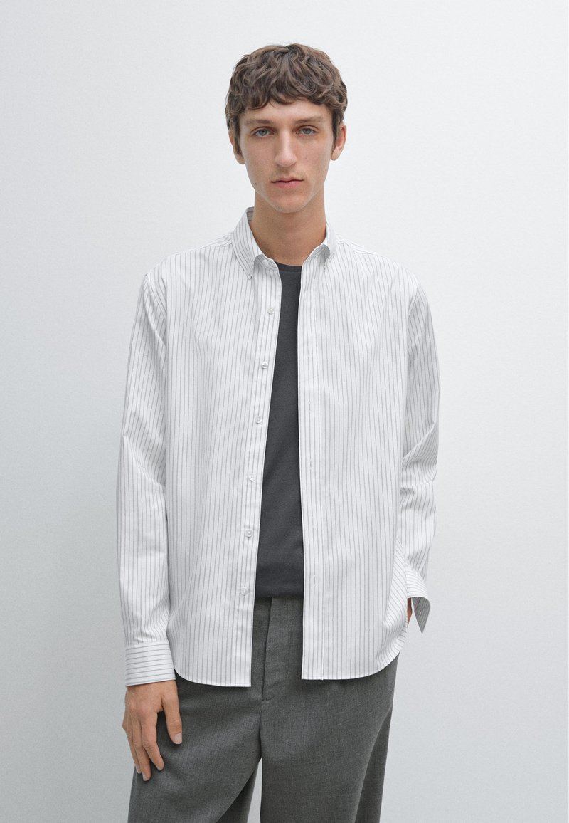 Camicia a righe bianca con bottoni, colletto, e orlo dritto; indossata sopra una maglietta grigia e abbinata a pantaloni grigi.