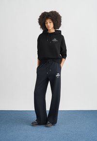 Μαύρο φούτερ με κουκούλα και κορδόνι καθώς και λογότυπο μπροστά, σε συνδυασμό με φαρδιά μπλε σκούρα φούτερ παντελόνια με λογότυπο, και τα δύο από απαλό ύφασμα.