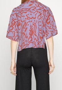 Kortärmad, croppad skjorta i lila tyg med ett rött flytande abstrakt mönster. Rak fåll med lös passform, inga synliga stängningar.