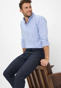 Chemise boutonnée bleu clair avec une poche poitrine, manches retroussées, associée à un pantalon foncé texturé, assis sur une chaise en bois.