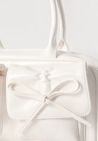 White faux leather handbag with a structured design, featuring a bow detail on a front pocket and two sturdy handles.