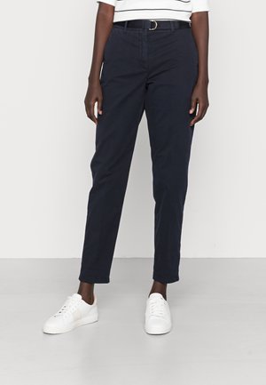 Person iført marineblå bukser med sort bælte, hvide sneakers og en hvid top med striber, stående på en lysegrå gulv mod en hvid væg.