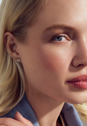 Guldørering i hjerteform prydet med små klare ædelsten, med en poleret finish. Nærbillede af øre og ansigtstræk.