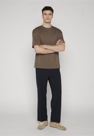 Młody mężczyzna stoi z założonymi rękami, ubrany w brązową koszulę z krótkim rękawem, czarne spodnie i beżowe trampki na jednolitym szarym tle.