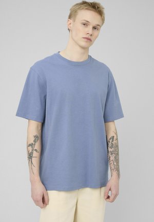 Jeune homme aux cheveux blonds courts portant un t-shirt bleu et un pantalon beige, montrant des tatouages sur les deux avant-bras, debout devant un fond uni.