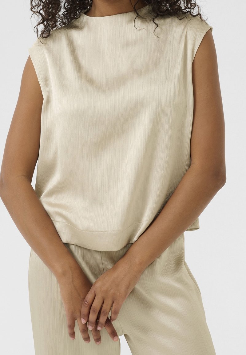 Femme portant un haut sans manches beige clair texturé et un pantalon assorti, les mains doucement jointes à la taille.