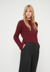 Burgundski krojeni pulover iz puhastega materiala z dolgimi, razširjenimi rokavi, v kombinaciji z visokim pasom in temno karirastimi hlačami s stranskimi žepi.