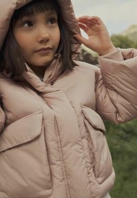 Puffjacke in hellrosa, mit hohem Kragen, großen Fronttaschen und einer glatten, glänzenden Textur. Kapuze teilweise zurückgezogen.