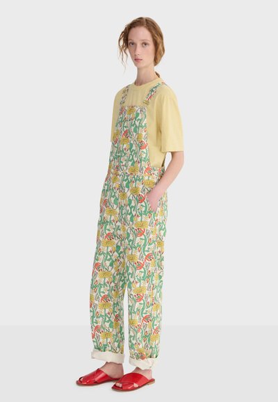 Femme portant une chemise jaune à manches courtes, une salopette à motifs floraux avec poches, et des sandales rouges ouvertes, debout devant un fond uni.