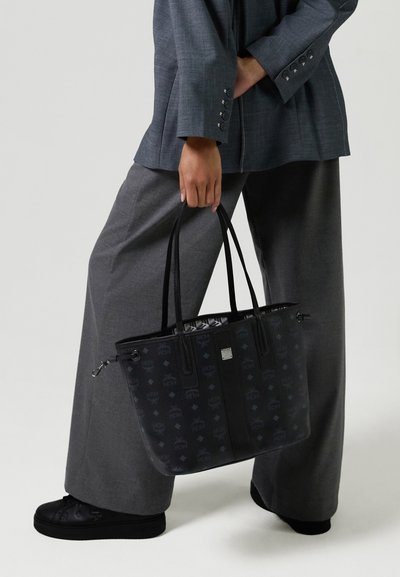 Personne portant un pantalon large gris et un blazer, tenant un sac fourre-tout noir à motif avec de longues poignées, des baskets noires visibles.