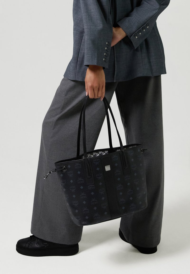 Persona in pantaloni ampi grigi e blazer che tiene una borsa a tracolla nera con fantasie e manici lunghi, sneaker nere in vista.