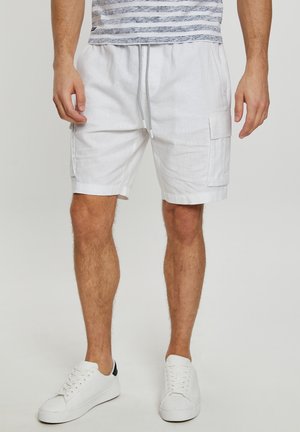Pantalones cortos cargo de lino blanco con bolsillos laterales, cintura elástica y cordón. Combinados con zapatillas blancas con un acento negro en el talón.