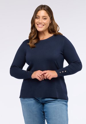 Lächelnde Frau mit welligem braunem Haar, die einen navyblauen Pullover mit goldenen Knöpfen an den Manschetten und hellblauen Jeans trägt, stehend mit gefalteten Händen.