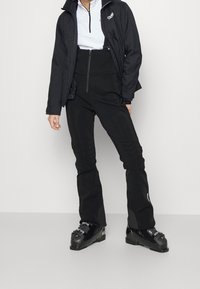 Person mit schwarzen Skihosen, schwarzen Skischuhen, einer schwarzen Jacke und einem weißen Oberteil mit Reißverschluss vor einem schlichten Hintergrund.