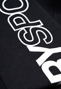 Tela negra con texto blanco en negrita. La textura parece suave y ligeramente elástica, con un diseño gráfico moderno.