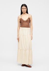 Γυναίκα με μακριά ίσια μαλλιά που φορά διαφανή γυαλιά, ένα καφέ τοπ χωρίς μανίκια με βολάν, μια μπεζ φούστα μακριά με βολάν και καφέ παντόφλες, όρθια.