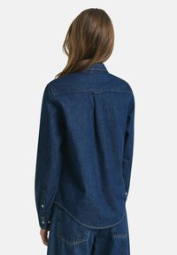 Woman with long brown hair wears a dark blue denim shirt and matching jeans, shown from the back against a plain light background.
