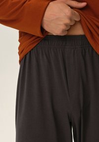 Brązowa bluzka z długim rękawem opadająca na ciemnoszare, lekkie, elastyczne spodnie z pasem w talii. Widoczna tekstura materiału jest gładka, z minimalnymi szwami i bez wzorów.