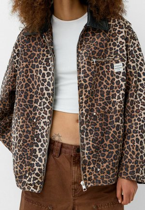 Persoon die een luipaardprint jas draagt over een witte crop top en een bruine broek, met een kleine zonnetatoeage op de onderbuik zichtbaar.