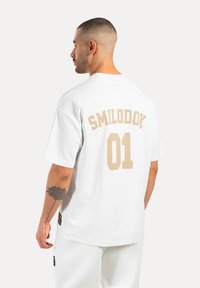 Witte katoenen t-shirt met korte mouwen, voorzien van beige geprinte tekst "SMILODOK" en "01" op de achterkant, ontspannen pasvorm, minimalistisch ontwerp.