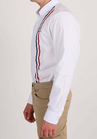 Homme portant une chemise blanche à boutons avec des rayures verticales rouges, blanches et bleues et un pantalon beige, vu de côté.